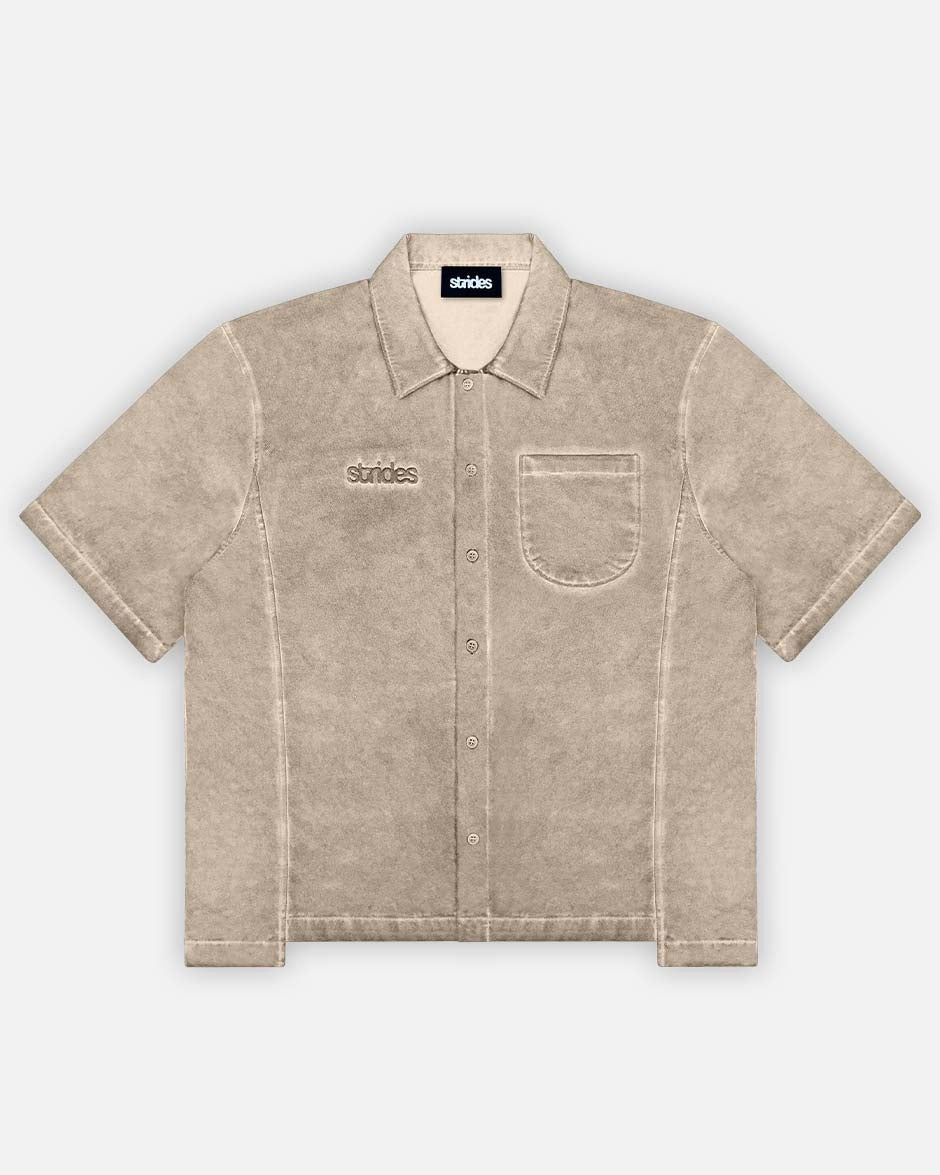 A beige, short-sleeved button-up shirt with a collar and front pocket, displaying a subtle oil washed texture and brand logo embroidery.