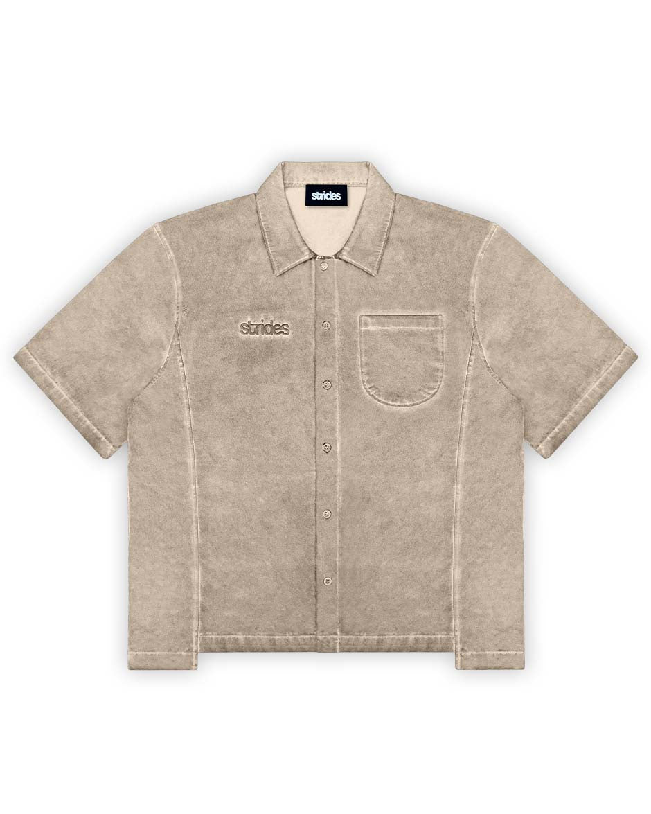 A beige, short-sleeved button-up shirt with a collar and front pocket, displaying a subtle oil washed texture and brand logo embroidery.