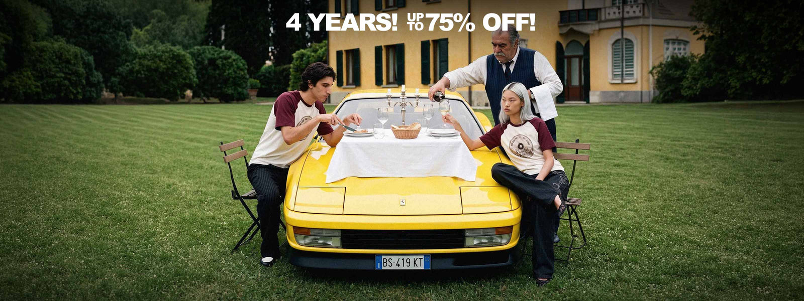 Two people in Strides raglan longsleeves and baggy trousers eating on the bonnet of a yellow Ferrari Testarossa in front of a yellow villa, while a waiter pours wine